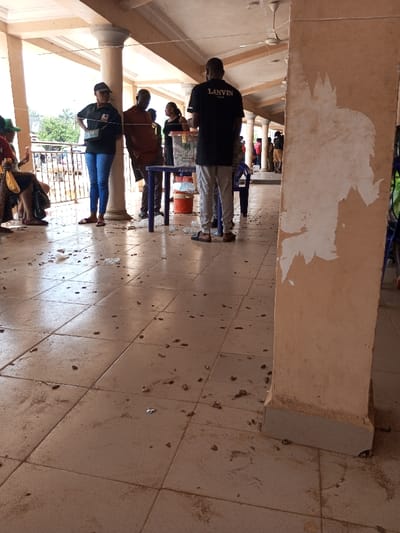 This image depicts a bustling, semi-enclosed public space in Ajalli, Nigeria, characterized by a tiled floor heavily soiled with dirt, scattered debris resembling peanut shells, and small pieces of plastic. The scene is illuminated by strong natural light entering from an open side of the structure.
In the foreground on the right, a large, rectangular pillar with peeling light-colored material, revealing uneven white patches, stands prominently. Towards the center of the frame, a group of individuals is gathered. A male subject, seen from behind, wears a black t-shirt with "LANVIN PARIS" text and light trousers, and stands facing a small, blue plastic table. On this table, a transparent container, an orange bucket, and other indistinct items are present. To the left of this individual, a female subject in a black shirt and blue jeans stands with her hands on her hips. Adjacent to her, another male subject in a brown shirt leans against a round, off-white pillar.
Further in the background, additional individuals are visible, some seated, some standing, suggesting a communal gathering or queuing. The architectural elements include multiple round pillars and an overhead roof structure, from which white ceiling fans are suspended. Beyond the covered area, trees and distant structures can be seen, indicating an outdoor or semi-urban environment. The overall setting appears to be a functional public area, possibly a temporary station or communal gathering point, marked by its well-used condition and the presence of widespread litter.
