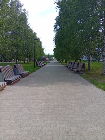 The image depicts a paved pedestrian walkway lined with benches and trees in Ruzaevka, Russia. The walkway is constructed from light-colored paving stones arranged in a regular pattern. On both sides of the walkway, there are benches made of wooden slats with metal frames, some of which are curved to form a continuous seating area. Tall green trees with dense foliage flank the walkway, providing shade. Several modern black lampposts are visible along the path. In the background, a few buildings and a red, curved structure are partially visible. The sky is overcast with a bright, diffused light. There are no people immediately visible on the walkway in the foreground, though a distant figure is present on the right side of the path further down. The environment appears to be a well-maintained park or public area.