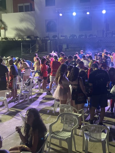 The image depicts an outdoor gathering in Blanca, Spain, possibly a festival or party, illuminated by stage lights. A crowd of young adults is visible, some standing and some seated on plastic chairs. In the foreground, a woman with long, dark curly hair is seated, holding a drink and looking to her right. She is wearing glasses and a dark sleeveless top, with a gold watch on her left wrist. Behind her, other individuals are present, and further back, a stage setup with lighting equipment is visible. The environment appears to be an open area with some architectural elements in the background, including buildings and railings. The overall atmosphere suggests a lively event taking place in the evening or night.