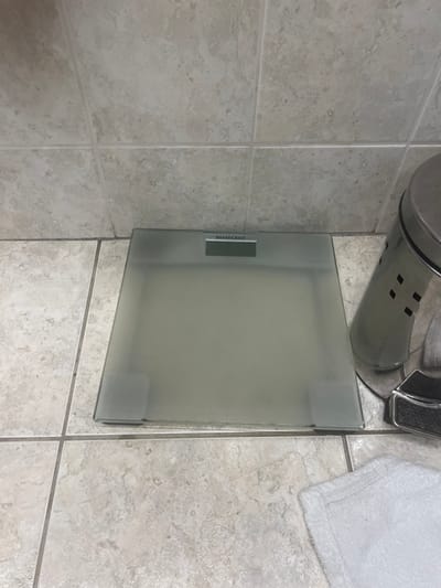 A top-down shot reveals a bathroom setting. A silver trash can sits adjacent to a frosted glass scale with a visible "Silvercrest" logo. A white towel is partially visible, occupying the top right corner. The floor is tiled in a neutral, speckled pattern, with grout lines clearly delineating each tile. The scale and trash can appear to be positioned in a corner space.