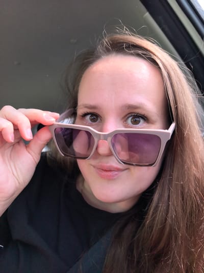 A close-up, eye-level shot shows a young woman holding light pink sunglasses up to her face, obscuring part of her eyes. She is looking directly at the camera, with a slight smile. The woman has long, brown hair that falls around her face. She is wearing a black t-shirt. The background is blurred, suggesting she is in a car. The scene is indoors, with natural light filtering in. There is no indication of the city of Nestiukovo, Russia, in this image.