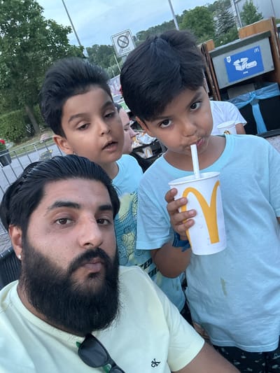 In a selfie taken at a McDonald's in Mainburg, Germany, a man with a beard and dark hair is in the foreground, wearing a light yellow t-shirt. Behind him, two young boys are visible. The boy on the right, wearing a light blue t-shirt, is drinking from a McDonald's cup with a straw. The boy on the left, also in a light blue t-shirt, looks towards the camera. In the background, there are trees, a sign indicating a "ZONE" with a prohibition symbol, and a recycling bin with McDonald's branding. The overall environment appears to be outdoors, possibly a parking lot or seating area of the fast-food establishment.