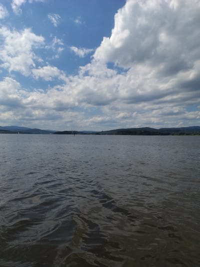 The image depicts a wide, dark body of water with gentle ripples under a cloudy blue sky. The horizon is lined with rolling green hills covered in trees. Several distant sailboats and a windsurfer are visible on the water, suggesting recreational activity. The overall scene conveys a sense of calm and open space, characteristic of a lake or large reservoir. It's plausible this is part of the Lipno Reservoir, a large artificial lake located in the South Bohemian Region of the Czech Republic, near the town of Bližná, which is situated close to the reservoir.