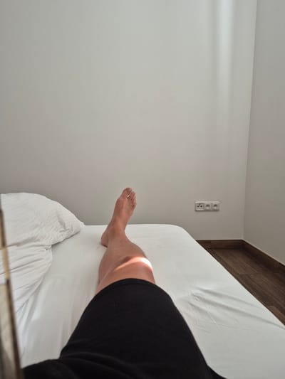 The image depicts a person's legs lying on a white-sheeted bed in a room. The bed is positioned in the lower half of the frame, with a white pillow visible on the left. The person is wearing black shorts. A shaft of sunlight illuminates the person's thigh. The background consists of a plain, light-colored wall with three electrical outlets mounted on it. The wall meets a wooden floor at the bottom right corner. The overall scene suggests a domestic or hotel room setting, likely in Poděbrady, Czechia, given the presence of European-style electrical outlets and the context provided.