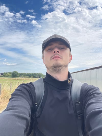 The image is a selfie taken outdoors on a partly cloudy day. A person with light-colored hair and a light beard is wearing a dark turtleneck shirt, a dark baseball cap, and a grey backpack. The person is looking directly at the camera. In the background, there is a grassy field with a fence, trees, and a corrugated metal building. The setting appears to be a rural or semi-rural area. While the image is described as being in Diepenbeek, Belgium, there are no specific visual cues within the image itself to confirm this location.
