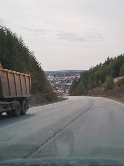 The image shows a road curving towards the city of Doksha, Russia. A large dump truck is traveling on the road in the same direction as the camera. Pine trees line both sides of the road, and the city of Doksha is visible in the distance. The sky is overcast.