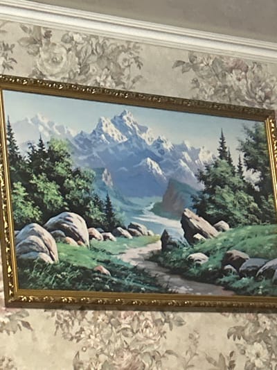 A framed landscape painting is displayed on a wall in Dmitriyevskaya, Russia. The painting features an ornate, gold-colored frame with decorative molding. The artwork depicts a mountainous scene under a light blue sky, with multiple snow-capped peaks in the background. A light blue river or stream winds through a valley in the midground, flanked by dense green coniferous trees and rocky slopes. In the foreground, a dirt path curves from the lower left towards the center, surrounded by numerous large, smooth, light-colored boulders and green grass. The wall behind the painting is covered with floral wallpaper, displaying a pattern of large, muted, light-toned flowers and leaves on a pale background. A white crown molding is visible along the top edge of the wall, bordering a white ceiling. No people or active subjects are present in the immediate environment.