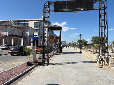 The image depicts a sunlit promenade in Năvodari, Romania, near the Black Sea coast. In the foreground, a wide, paved walkway is bordered by a street on the left and a low stone wall with landscaping on the right. The street features parked cars, including a dark sedan with a license plate visible (CT 01 GDK), and a bus stop sign.
Dominating the left side of the promenade is a large, dark metal structure, possibly an archway or part of an overhead display. To its right stands a small, wooden kiosk with a brown, pitched roof. Beyond this, the promenade extends into the distance, with several people walking.
The environment suggests a coastal urban setting. Buildings with multiple floors and balconies are visible in the background. Street lamps line the right side of the promenade, and various trees and shrubs are planted along the stone wall. The sky is clear blue with scattered white clouds. A portable toilet is positioned near the stone wall on the right. The overall scene is one of a public walkway on a bright, clear day.