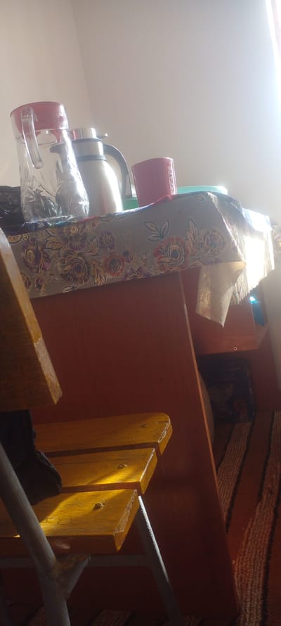 The image shows a cluttered indoor scene. There are several mugs, a thermos, and a decorative fabric item with a floral pattern stacked on top of a wooden cabinet. The cabinet itself is brown, and a portion of a yellow wooden chair is visible in the foreground. The background includes a striped rug. The location is unknown.