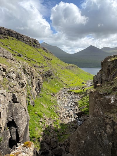 The image depicts a rugged, mountainous landscape in the vicinity of Haldórsvík, Faroe Islands. The scene is dominated by steep, verdant slopes interspersed with rocky outcrops and a narrow, rocky ravine. A small stream flows through the ravine, disappearing into the distance. In the mid-ground, a solitary, small, dark-roofed building with stone walls is visible on a grassy incline. Further in the distance, a body of water, likely a fjord or bay, is visible, with conical mountains rising beyond it under a partly cloudy sky. The lighting suggests it is daytime, with patches of sunlight illuminating the landscape. The overall impression is one of a remote and wild natural environment.