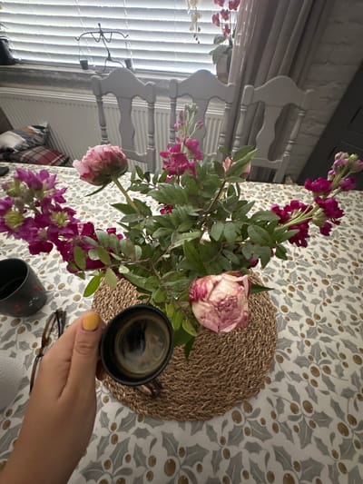 An overhead shot shows a hand with yellow nail polish holding a small black cup of coffee. The cup is positioned on a woven placemat, which rests on a tablecloth with a holly and berry pattern. In the background, a bouquet of pink and purple flowers is arranged in a basket. To the left, a pair of glasses and a dark mug with a floral design are visible. The scene appears to be an indoor setting, possibly a dining room, with chairs and a radiator visible in the background. The overall ambiance suggests a quiet moment of relaxation. The image was taken in Macclesfield, United Kingdom.