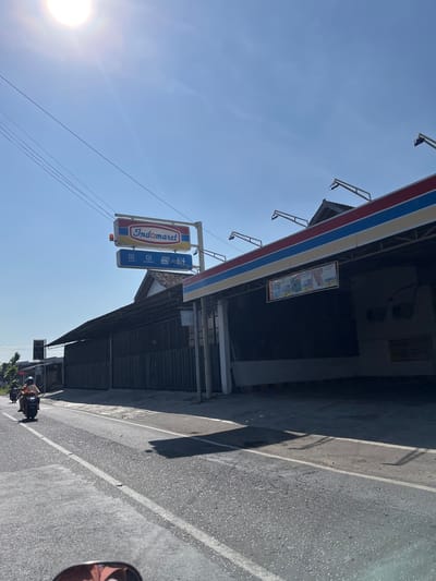The image captures a street scene in Purworejo, Indonesia, under a bright, sunny sky. The foreground shows a portion of a road with white lane markings, and what appears to be the handlebars and mirror of a vehicle from which the photo is taken. Further down the road, a person is riding a scooter. To the right of the road is a building with a red, white, and blue striped awning. A prominent sign for "Indomaret" is visible above the building. This Indomaret store has a covered, open-air structure extending from it, with a dark, corrugated metal wall behind. Several promotional banners are hanging under the awning. The building is situated along a paved road, with power lines running overhead.