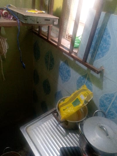 A kitchen scene shows a stainless steel sink. Above the sink, yellow and gold cookware contains a yellow packet. A covered pot sits to the right. A barred window allows light to stream in. The wall is tiled with a blue and white leaf pattern. A shelf holds a metal object with a blue cord. A coil of rope hangs nearby.