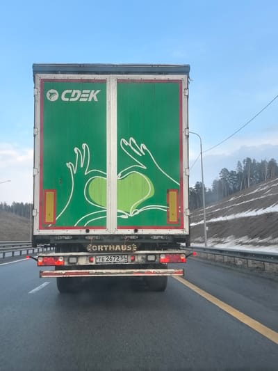 The image depicts the rear of a green CDEK delivery truck with a white logo and graphic of two hands forming a heart. The truck is on an asphalt highway with a yellow lane marker. The license plate reads "TE 2672 54 RUS". The ORTHAUS brand is visible above the license plate. The environment includes a hillside covered with snow and trees. This image was taken near Сапуголи, Russia.