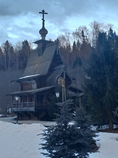 The image shows the Church of St. Sergius of Radonezh in Vzglyadnevo, Russia. The wooden church stands surrounded by trees and a snow-covered ground. A cross tops the structure. A small evergreen tree is present in the foreground. The sky is overcast.