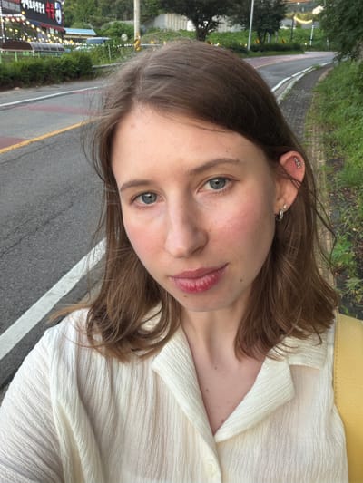 A young woman with brown hair and blue eyes is taking a selfie outdoors in front of a road in Pocheon-si, South Korea. She is wearing a cream-colored collared shirt and a yellow bag is visible over her shoulder. She has multiple ear piercings. The road is paved with yellow and white lines, and there is a sidewalk with paving stones and grass on the right side. In the background, there are trees, a building with neon lights, and a street lamp. The lighting suggests it is dusk.