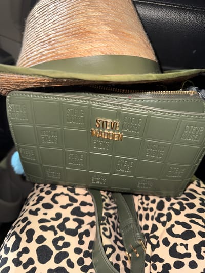 A close-up, top-down view captures a straw fedora hat with a green band resting on a green textured wallet. The wallet, which features repeated "STEVE MADDEN" embossing in a grid pattern, is placed on top of a pair of leopard-print pants. A green strap from a bag is visible on the right side of the frame, partially obscuring the pants. The scene is indoors, and the lighting suggests it was taken during the day.