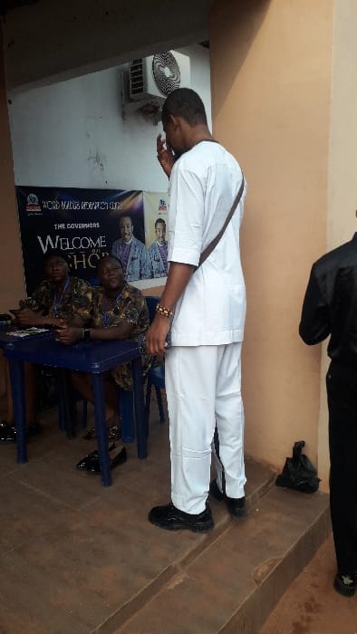 The image captures an indoor scene, likely a public hall or reception area in Awada, Nigeria. In the foreground, a man stands, dressed in a full white traditional ensemble consisting of a long-sleeved tunic and trousers, paired with black dress shoes. A brown strap of a shoulder bag is visible across his chest. He wears a beaded bracelet on his left wrist. The man is bowing his head downwards, with his hands positioned around his waist, suggesting a gesture of respect, prayer, or humility.

Behind the standing man, two individuals are seated at a table on blue plastic chairs. They are wearing patterned dark clothing, possibly traditional African print attire, and appear to be attending to something on the table. Their faces are partially visible.

To the left of the standing man, an air conditioning unit is mounted on a light-colored wall. A large banner or poster is prominently displayed behind the seated individuals and partially behind the standing man. The banner features images of men in traditional wear and several lines of text. Clearly visible are "WELCOME" in large white letters, and below it, "THE GOVERNORS" and "CHO". Above "WELCOME", partially visible text reads "WOHD MAKE RES...ON".

The floor appears to be a light brown tiled or concrete surface. The walls are a mix of light brown/beige and white. The overall lighting suggests it is daytime. Another person's legs and shoes in black attire are partially visible in the upper right background.