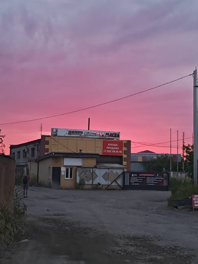 In the city of Vasilkovo, Russia, a pink and purple sky, likely from a sunset, dominates the scene. In the foreground, a dirt road leads towards a cluster of buildings. A person with a backpack is walking away from the camera on the left side of the road. The buildings appear to be industrial or commercial, with signs indicating "CENTER OF SPARES" and "RENT AND SALE" with a phone number. A metal gate with an advertisement is visible on the right side of the frame. Electrical wires are strung across the sky.
