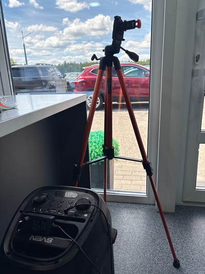 The image shows a tripod with an orange pole extended diagonally across the frame. A black speaker with the brand name "SVEN" visible is positioned in the foreground on the right side. The speaker has various controls and ports, including buttons for "MIC," "MICRO SD," "AUX," "USB," and "POWER," as well as knobs for "VOL" and "MIC 2." A power adapter is plugged into the speaker.
The background suggests an indoor setting with a large window looking out onto a street scene. A red car is visible through the window, along with a paved area and some green foliage. The sky with clouds is also visible through the window, indicating it is daytime. The overall lighting suggests a bright day. There are no people visible in the image.
The text "PRO 1800" is visible on one of the tripod's legs, likely indicating the model number. The setting appears to be a public space or a store, possibly an electronics store, given the presence of the speaker and tripod. The orientation of the image is landscape and it is likely a photograph.