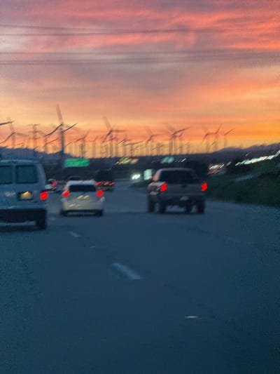 This image captures a highway scene at dusk or dawn in the Palm Springs area of the United States. The sky dominates the upper portion of the frame, painted in vibrant hues of orange, pink, and soft purple, indicating either a sunset or sunrise. Several dark power lines stretch horizontally across the upper third of the sky.

In the mid-ground, a vast wind farm is visible on distant hills and mountains, featuring numerous wind turbines with their blades silhouetted against the colorful sky. The turbines appear in various states of motion blur, suggesting they are spinning. The landscape beyond the highway appears arid and mountainous.

The lower half of the image shows a multi-lane highway, where several vehicles are traveling. The scene is somewhat blurry, especially the moving cars, implying the photo was taken from a moving vehicle. In the foreground on the left, the rear of a light-colored van is partially visible. Further down the road in the center, a light-colored sedan with illuminated red taillights is seen. To its right, a dark-colored pickup truck also displays red taillights. White lane markings are faintly visible on the dark asphalt of the road.

The overall impression is one of movement and travel through a landscape characterized by both natural beauty (the sunset/sunrise and mountains) and modern energy infrastructure (the wind farm). No explicit text is clearly visible in the image.