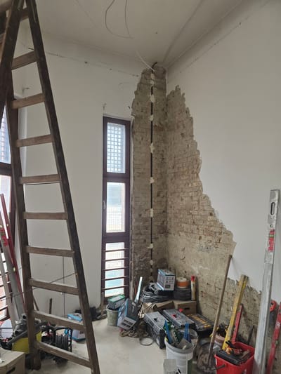 The image depicts an interior room in Nyíregyháza, Hungary, undergoing active renovation or deconstruction. The scene is characterized by exposed brickwork, construction materials, and tools.
On the right side, a substantial portion of the wall plaster has been removed, revealing underlying red brick and mortar with an irregular, jagged edge where it meets the remaining white plaster. A vertical black conduit or cable is affixed to the exposed brick corner with multiple white fasteners.
The ceiling is white with visible surface imperfections and several white electrical wires routed across it, some secured, others loosely hanging. The floor is a light, dusty surface, likely concrete or screed, covered with debris and equipment.
In the left foreground, a tall, dark brown wooden A-frame ladder stands, partially obscuring the view of the wall behind it. Two tall, narrow windows with dark frames and internal grids are visible in the background, allowing natural light into the room.
Various construction tools and materials are scattered across the floor, particularly on the right side of the room. These include: a red plastic bucket filled with hand tools (trowels, spatulas); a long-handled shovel; a yellow spirit level and a silver spirit level leaning against walls; a yellow and black toolbox; coiled electrical cables; cardboard boxes; a white bucket containing tools; stacks of circular objects; a red pressurized canister; and a yellow pressure washer or similar device with a hose reel. A large coil of rope is also present on the floor. No individuals are visible.