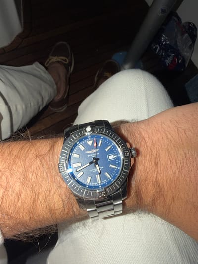 The image shows a person's left arm wearing a blue Breitling Chronometre Avenger wristwatch. The arm is covered in dark hair. The person is wearing white pants. In the background, there is a wooden floor, a dark blue bag, and a metal pole. The scene is indoors and dimly lit. There is no identifiable information that can place this image in Tønsberg, Norway.