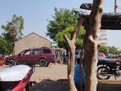 A red station wagon is parked on a dirt road in front of a building with a sign reading "RIBADU." Two large trees are on the right side of the frame. A man wearing a blue shirt is sitting next to the trees. There are a few other vehicles visible, including a white pickup truck. The scene is in Tongo, Nigeria, with the coordinates 10.664 latitude and 11.364 longitude. The area has a dirt road, buildings, and trees.