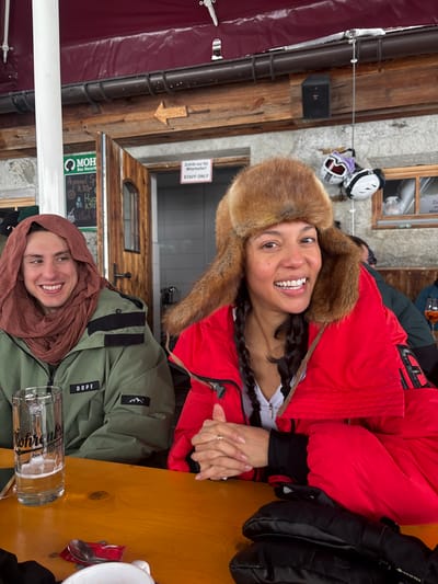 The image captures two individuals seated at a light brown wooden outdoor table beneath a red awning, likely at an establishment in St. Christoph, Austria.
On the left, a male subject with light skin and short dark hair, wearing a reddish-brown scarf or hood and a green winter jacket displaying "DOPE" and a mountain logo, smiles towards the right, revealing his teeth. A large glass beer mug, branded "Fohrenburg das Vorarlberger Bier" and containing a light-colored liquid, is on the table in front of him.
On the right, a female subject with medium skin tone, wearing a red winter jacket, a white top, and a brown fur ushanka-style hat, smiles directly at the viewer, also revealing her teeth. Her dark hair is styled in two braids. Her hands are clasped on the table, with a ring visible on her left ring finger. A pair of black mittens or gloves rests on the table to her right.
The background consists of a building with a stone foundation and wooden walls. A wooden door with an arched window is visible, featuring a white sign that reads "Zutritt nur für Mitarbeiter! STAFF ONLY". Above the door, a wooden arrow points left. To the left of the door, a green sign for "MOH das Vorarlberger Original" displays "Aperol Spritz 8,50.-". A small black speaker is mounted on the wooden wall, and ski goggles (white and purple) are hanging near a window. Blurry figures are discernible further in the background. A white support pillar, a metal gutter, a small white dish, a red paper napkin, and a spoon are also visible on the table.