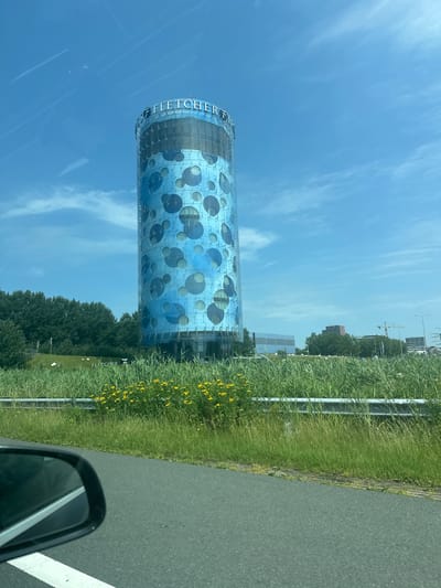 The image shows a cylindrical building with a blue facade featuring large circular patterns, located in Ouderkerk aan de Amstel, Netherlands. The building has "FLETCHER HOTELS" signage at the top. In the foreground, there is a road with a white line marking and a car's side mirror. A grassy embankment with yellow flowers separates the road from the building, and beyond the building, trees and other structures are visible under a blue sky with scattered clouds.