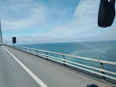 This is a landscape photograph taken from a vehicle moving across a bridge in Denmark. The perspective is from the driver's or passenger's side of the vehicle, looking forward and slightly to the right. The road is asphalt with a white dashed line marking the lane. A metal railing runs along the side of the bridge, separating the road from the vast expanse of blue water. In the distance, on the horizon, are several wind turbines and a small yellow buoy. The sky is a clear blue with scattered white clouds. A dark, blurry object, likely part of the vehicle's interior such as a rearview mirror or a dashcam, is visible in the upper right corner of the frame. The overall impression is one of a modern infrastructure crossing a body of water under a bright, partly cloudy sky.
