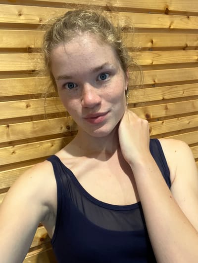 A fair-skinned woman with blonde hair and blue eyes is lying on a wooden surface, possibly a sauna wall, and looking at the camera. She is wearing a dark blue swimsuit. Her left arm is raised and bent, with her hand near her neck. The background is a wooden wall made of narrow, light-colored planks with small dark knots. The location in the city of Pobrdje, Montenegro cannot be verified from the image.