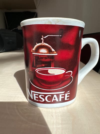 A close-up, eye-level, indoor shot shows a white mug with a red design featuring a vintage coffee grinder and a cup of coffee. The word "NESCAFÉ" is prominently displayed in white letters on the mug's surface. The mug is positioned on a light-colored, textured surface, possibly a table. Sunlight casts a shadow on the surface to the left of the mug, creating a stark contrast and highlighting the object. In the blurred background, dark, indistinct shapes suggest a domestic or office environment. There is no discernible location in Nantes, France, evident in the image.