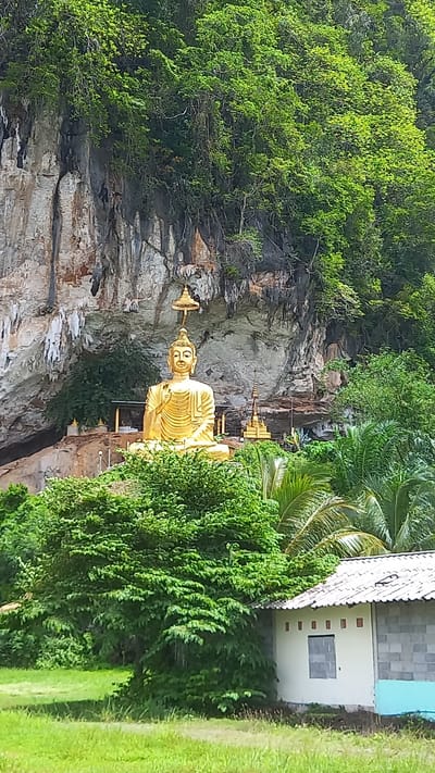A landscape scene features a golden Buddha statue positioned in front of a rocky cliff. The statue, centered in the frame, is a prominent figure, contrasting with the rock formation behind it. Lush greenery surrounds the statue. A building with a gray tiled roof and blue walls is on the right side of the frame. A grassy area is visible in the lower right corner. Based on available information, the scene is in Thap Put, Thailand.