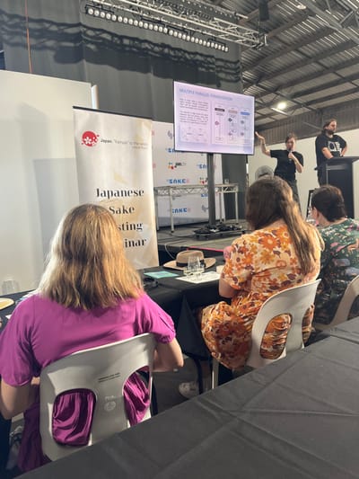 The image shows a Japanese Sake Tasting Seminar in Hamilton, Australia, located at the coordinates -27.438 latitude and 153.078 longitude. Three people are seated at a table, listening to a speaker who is standing on a stage in the background. The speaker, dressed in a black shirt and black pants, is pointing at a presentation on a screen with a title that reads "Multiple Parallel Fermentation". Behind the speaker, on the stage, a logo for "JAMS.TV" can be seen. The person on the left, a female, is sitting in a white chair wearing a magenta t-shirt. Another female, seated to the right, wears a floral dress. A third person, a female, is seated to the right of the second female.  The table is covered with black fabric. There are white chairs in front of the table. A straw hat lies on the table.  Several glasses of liquid are set out on the table in front of the people. There is a sign on the stage that reads "Japanese Sake Tasting Seminar". Another sign behind the table reads "Japan "Kampai" to the world Sake & Shochu".  The room is well lit and has a high ceiling, with exposed beams and pipes.  It appears to be a warehouse-type space, possibly an event venue.