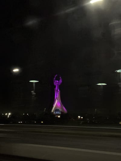 The image depicts a nighttime scene in Jarbashi, Uzbekistan, with a prominent illuminated statue in the center. The statue, which appears to be of a winged figure, is lit with a vibrant purple hue and has a distinct triangular base. To the left and right of the central statue, two other illuminated structures are visible, resembling large mushroom-like or disc-shaped lights on tall poles. These are also lit, though their colors are less distinct and appear greenish or white. In the foreground, a dark road or highway is visible, suggesting the photo was taken from a moving vehicle. The background is a dark, indistinct cityscape with scattered lights, and the overall atmosphere is that of urban night illumination. The image quality is somewhat degraded, with reflections and motion blur present, consistent with a photograph taken through a window at night.