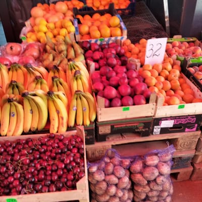A market stall in Cupcini, Moldova, displays an abundant selection of fresh produce. The foreground is dominated by a wooden crate overflowing with dark red cherries. To the right of the cherries, several netted bags are packed with what appear to be root vegetables, possibly beets or potatoes, with their earthy skins visible through the netting.
Moving further into the frame, various fruits are arranged in wooden crates and boxes. Bunches of bright yellow bananas are prominent, with some still showing their green stems. Next to them, a pile of red peaches or nectarines are neatly arranged, their smooth skins gleaming. In the background, a broader assortment of fruits is visible, including oranges and lemons, filling multiple crates. One crate prominently features a white sign with the number "32" handwritten in black. The overall impression is one of a vibrant and well-stocked produce market, showcasing the bounty of local agriculture. The scene is lit with natural light, contributing to the fresh and colorful appearance of the fruits and vegetables.