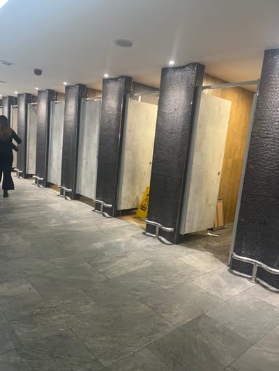 This image depicts an indoor public facility, most likely a restroom or changing area, characterized by a long row of individual stalls. The floor is laid with large, light grey, rectangular tiles. The partitions separating the stalls are dark, appearing to be covered in small, possibly mosaic, tiles, and are supported by silver-colored fixtures. The stall doors are a solid, light color, such as white or light grey. The ceiling is white and features multiple recessed spotlights, providing artificial illumination.
A yellow "wet floor" or "caution" sign is visible on the tiled floor in the mid-ground, positioned near the entrance to one of the stalls, suggesting recent cleaning or a potential hazard. Beyond the stalls, a warm-toned wall or panel, possibly wood, is discernible.
In the foreground, a person dressed in dark clothing is partially visible, walking away from the camera. The scene suggests a modern and functional environment. No specific time of day or outdoor weather conditions are apparent due to the indoor setting and artificial lighting. The only prominent visible text is the universal caution symbol (black triangle with an exclamation mark) on the yellow sign.
