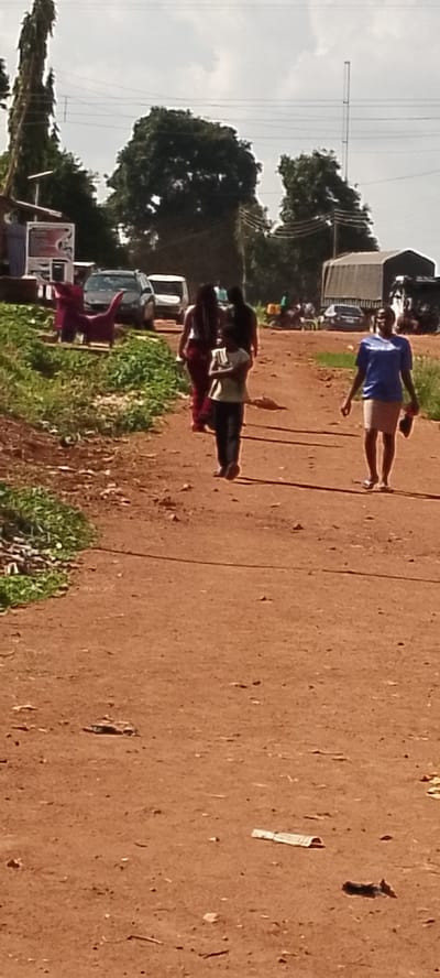 In the city of Ajio, Nigeria, a group of people walk along a dirt road. The road is a reddish-brown color, suggesting a dry, dusty environment. On the left side of the road, there is a patch of green grass and some debris. In the background, trees and buildings are visible. A tall transmission tower stands in the distance. There are several vehicles on the road, including cars and a truck covered with a tarp. A red, plastic chair shaped like a dinosaur is on the side of the road near a building. The sky is overcast.