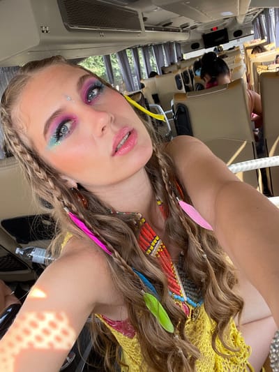 A woman with vibrant, colorful makeup poses inside what appears to be a bus. Her hair is styled with braids and adorned with feathers, and she wears a beaded necklace over a yellow, crocheted top. She has shimmering eye makeup with shades of pink, blue, and glitter, and a rhinestone is placed on her forehead. Passengers and seats can be seen in the background.