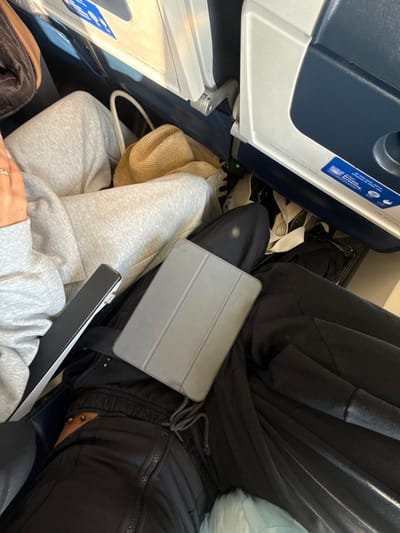 The scene depicts the interior of an airline cabin, consistent with an aircraft at or en route to/from Charles de Gaulle Airport (CDG) in Roissy-en-France, France. Two individuals are partially visible, seated in adjacent passenger seats. The person on the left wears light gray sweatpants and a matching long-sleeved top; a light brown straw hat rests on their lap. Their right hand, adorned with a ring, is visible. A black smartphone with a metallic edge is positioned near their left leg on an armrest. The person on the right, clad in black track pants featuring a drawstring and a zippered pocket, has a gray tablet device with a folding cover resting on their lap. A gold-colored beaded ankle bracelet is visible on their right ankle. The immediate environment includes blue and white seatbacks, part of a tray table mechanism, and various personal belongings such as a tan bag in the upper left and dark fabric items on the floor. A small green indicator light is visible near the floor. A partial airline safety instruction label on the seatback reads, "Fasten your seat belt while seated."