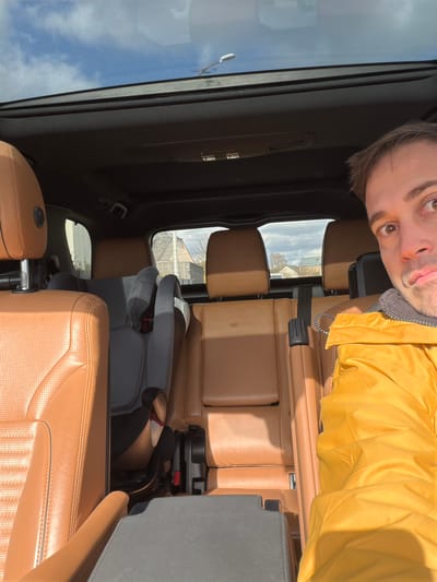 A selfie taken inside a vehicle reveals an adult male and the vehicle's interior. The male subject, positioned on the right of the frame, has light brown hair and brown eyes, wearing a yellow jacket over a grey garment, with a neutral expression. The vehicle's interior features tan/brown perforated leather seats across multiple rows. A grey child safety seat is installed in the center of the second row. A panoramic sunroof is visible overhead, showing a clear blue sky with scattered white clouds, indicating daylight. Through the rear window, residential buildings with pitched roofs and bare trees are visible, along with a distant white vehicle. The vehicle is located in Reckange-sur-Mess, Luxembourg.