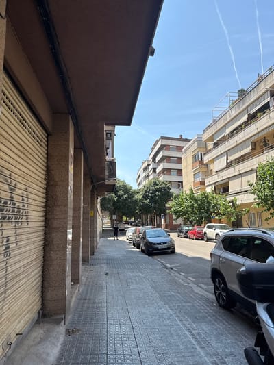 The image depicts a street scene in Vilanova i la Geltrú, Spain, on a sunny day. The foreground shows a sidewalk paved with textured tiles, running alongside a building with a large, closed metal shutter on the left. Several large concrete pillars support an overhang of the building. The street is lined with parked cars and trees. In the distance, more apartment buildings are visible, and contrails streak across the clear blue sky. A single person is walking on the sidewalk in the middle ground. The overall atmosphere is that of a typical urban environment.