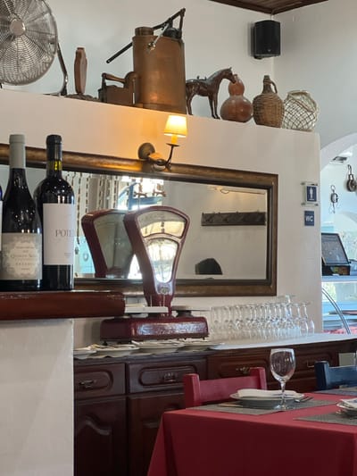 An interior view of an establishment in Coruche, Portugal, displays a dining area and a decorated counter space. In the foreground to the right, a table is set with a red tablecloth and a single place setting: a grey placemat, white plate, folded napkin, fork, knife, and an empty stemmed wine glass. A red wooden-framed chair is partially visible.
Behind the table, a dark wooden cabinet holds a stack of white plates and a prominent red vintage mechanical weighing scale with two dials. To its right, approximately twenty empty stemmed wine glasses are arranged. Above the cabinet, a large rectangular mirror with a dark wooden frame is mounted on a white wall, reflecting a dark coat rack and a blurred form resembling a person's head. A wall-mounted sconce with a yellow-lit lampshade is affixed above the mirror.
To the right of the mirror, two blue square signs on the white wall indicate facilities: an upper sign with male and female pictograms, and a lower one marked "WC." Further right, an arched doorway reveals an area with a display case and hanging objects.
On a wooden ledge in front of the mirror, two dark glass wine bottles are displayed, one partially labeled "QUINTA NOVA RESERVE" and the other "POTE." Above, a white shelf showcases various decorative objects: a large silver industrial fan, a copper manual pump, a dark vintage flat iron, a wooden decorative piece, a dark horse figurine, two gourd-shaped ceramic vessels, and two woven straw-covered bottles. In the upper right corner, a black speaker is mounted, and dark wooden ceiling beams are visible. The scene is brightly lit, with a reflection suggesting a person's presence elsewhere in the room.