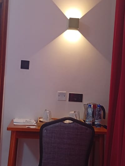 This image depicts a segment of an indoor room, identified as being in Murang'a, Kenya. The scene is dominated by a white wall, on which a cube-shaped wall sconce is centrally mounted, emitting light upwards and downwards. Below the light fixture, a black square-shaped object is affixed to the wall, followed by two white electrical outlets and a dark rectangular control panel. A light brown wooden table with angled legs is positioned against the wall. On the table's left side, a stack of white papers or a brochure is visible. To the right, an arrangement for beverages includes a black electric kettle, two plastic bottles of "Dulton" branded water, and two clear inverted glasses resting on a light wooden tray. The upper backrest of a dark grey fabric chair, featuring a black frame and metallic studs, is prominent in the immediate foreground, partially obscuring the table. A vertical section of a red curtain is visible along the far right edge of the frame. No individuals are present in the scene.