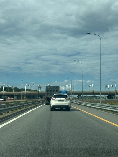 The image shows a highway in the city of Levashovo, Russia, under a cloudy sky. The perspective is from inside a moving vehicle, looking forward. In the foreground, the asphalt road is visible with a white lane line on the left and a yellow lane line on the right. A white SUV is driving in the right lane, in front of the camera vehicle. To the left of the SUV, a dark-colored truck is also on the highway. Beyond the vehicles, a large, multi-level overpass structure with distinctive curved supports dominates the middle ground. Streetlights line the highway on both sides. The background shows trees and more of the overpass structure. The overall scene depicts a typical highway environment with traffic under an overcast sky.