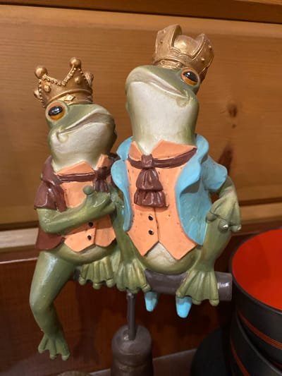 Two ceramic frog figurines, dressed in royal attire, are positioned on a branch. The frog on the left wears a brown vest and an orange undershirt with a brown bow tie. The frog on the right is adorned with a blue jacket over an orange vest and a brown bow tie. Both wear golden crowns. The background is a wooden wall. The scene appears to be indoors, possibly in a residential or commercial setting, with a portion of red and black bowls visible in the lower right corner. No specific details regarding the city of Ленинское, Russia, are present in the image.