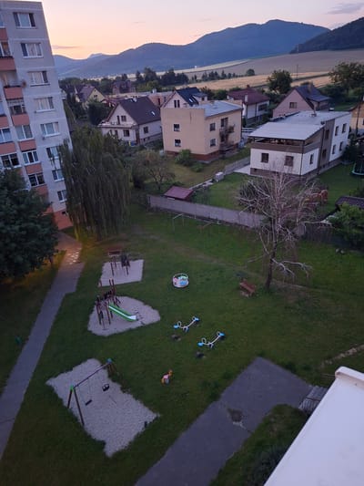 This image captures a residential area in Ilava, Slovakia, during twilight. A tall, multi-story apartment building dominates the left side of the frame. The foreground features a playground with swings, a slide, and two seesaws. A barren tree with intricate branches stands in the middle of a grassy area, with a wooden bench beneath it. In the background, several houses with pitched roofs are visible, nestled amongst trees. Rolling hills and fields, illuminated by the soft glow of the setting sun, form the backdrop, with a hazy mountain range in the far distance. The sky displays a gradient of soft pinks and blues, indicative of the transition from day to night. The scene appears peaceful and suburban.