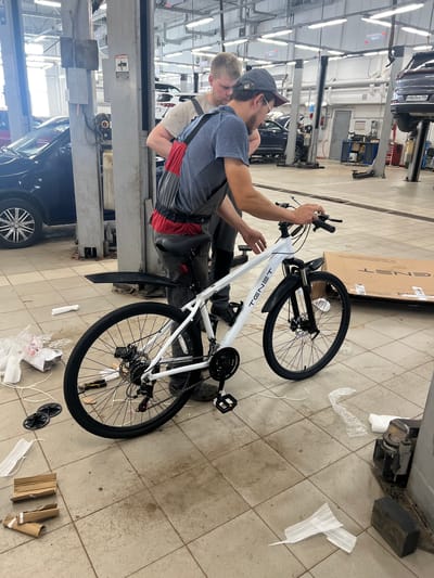 Two men are assembling a white bicycle in a brightly lit, tiled garage or workshop. The bicycle has black fenders and tires. Tools are scattered on the floor near the bike, including pliers and what appears to be a small wrench. Packaging materials, including cardboard and plastic wrap, are also present. In the background, several cars are visible, including one on a hydraulic lift. One man, wearing a cap and glasses, is holding the handlebars of the bicycle, while the other man stands behind him. The environment suggests a commercial or industrial setting, possibly a car repair shop or a bicycle retail store. The location is identified as Люк, Russia.