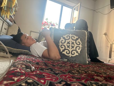 A young man is lying on a rug in a room, resting his head on a pillow. He is wearing a white t-shirt. Behind him, a person in a dark hooded garment is seated, holding a patterned cushion. A window is open behind them, letting in bright daylight. Red flowers are visible in pots on the windowsill. The room appears to be a bedroom, with a dresser and a bed or mat on the floor. The rug has an intricate red and black pattern. A white cord, possibly from earphones, is visible on the rug. The scene suggests a relaxed, indoor setting during the daytime.